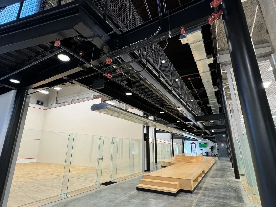 Interior of a modern sports facility, featuring squash courts, elevated walkway, and exposed industrial infrastructure.
