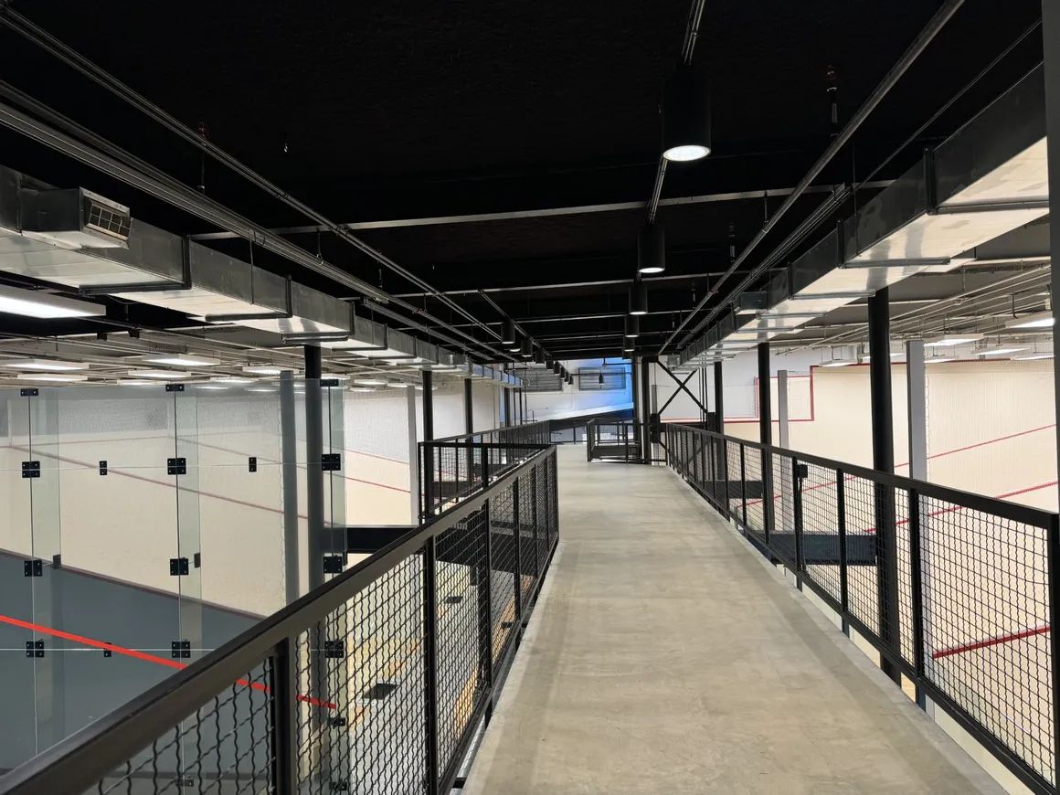 Interior view of a walkway overlooking multiple squash courts. Black railings, gray walkway, and clear glass walls are visible.