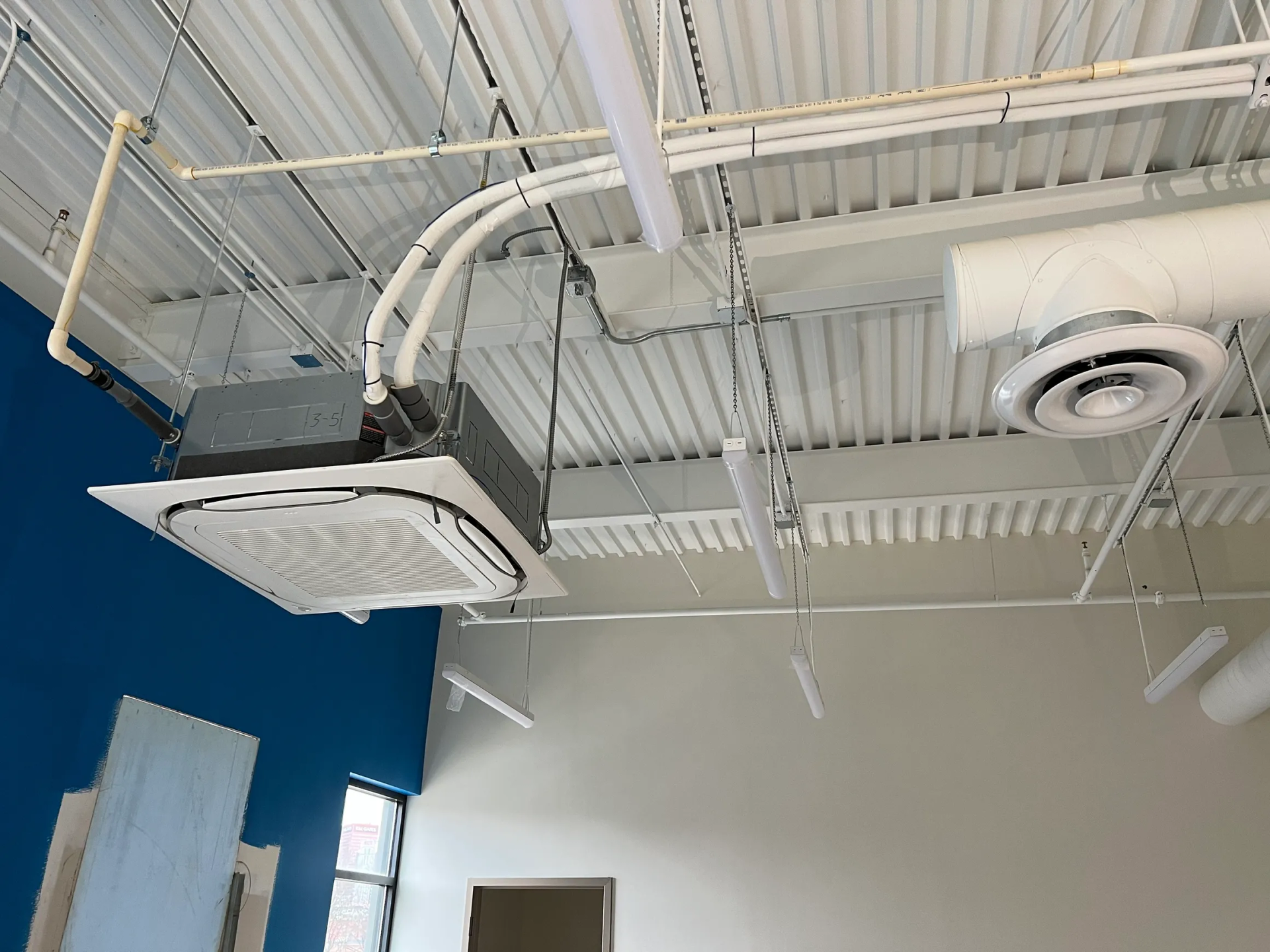 Ceiling-mounted HVAC unit with ductwork and lighting in a commercial space, white and gray with blue accent wall.