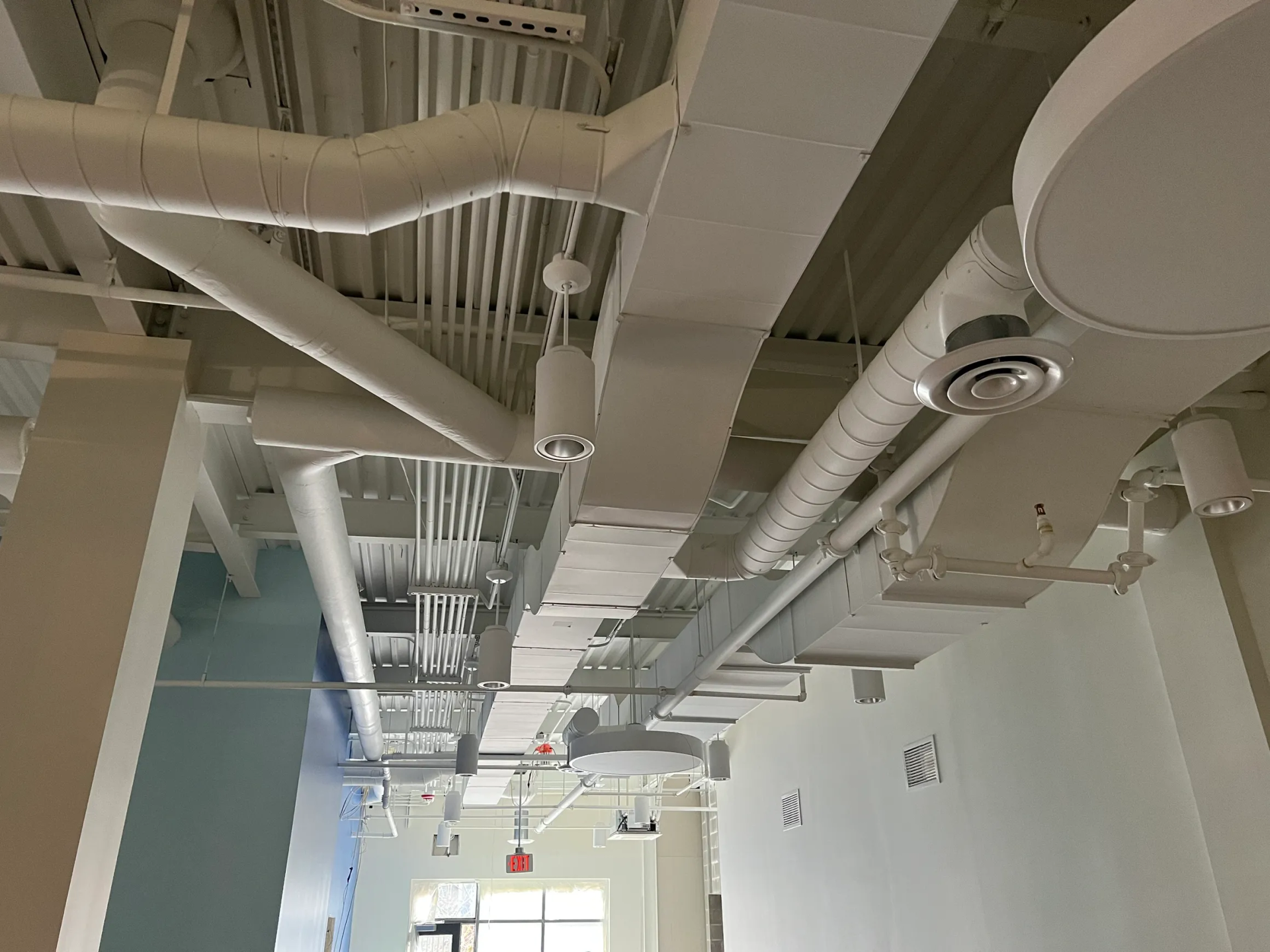 White ceiling with exposed pipes, ducts, and lighting fixtures in a commercial space.