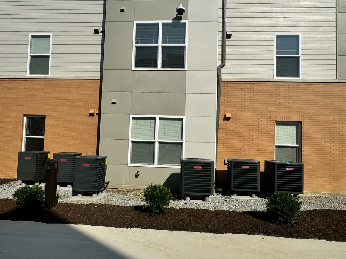 Exterior view of a building with air conditioning units lined up on the ground in front of a brick and panel facade.