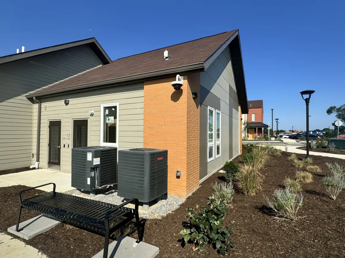 Exterior view of a modern building with brown and tan siding, HVAC units, a bench, and a brown roof.