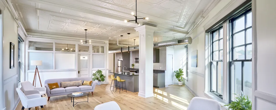 Spacious, bright living area with kitchen, large windows, white walls, and wood floors.