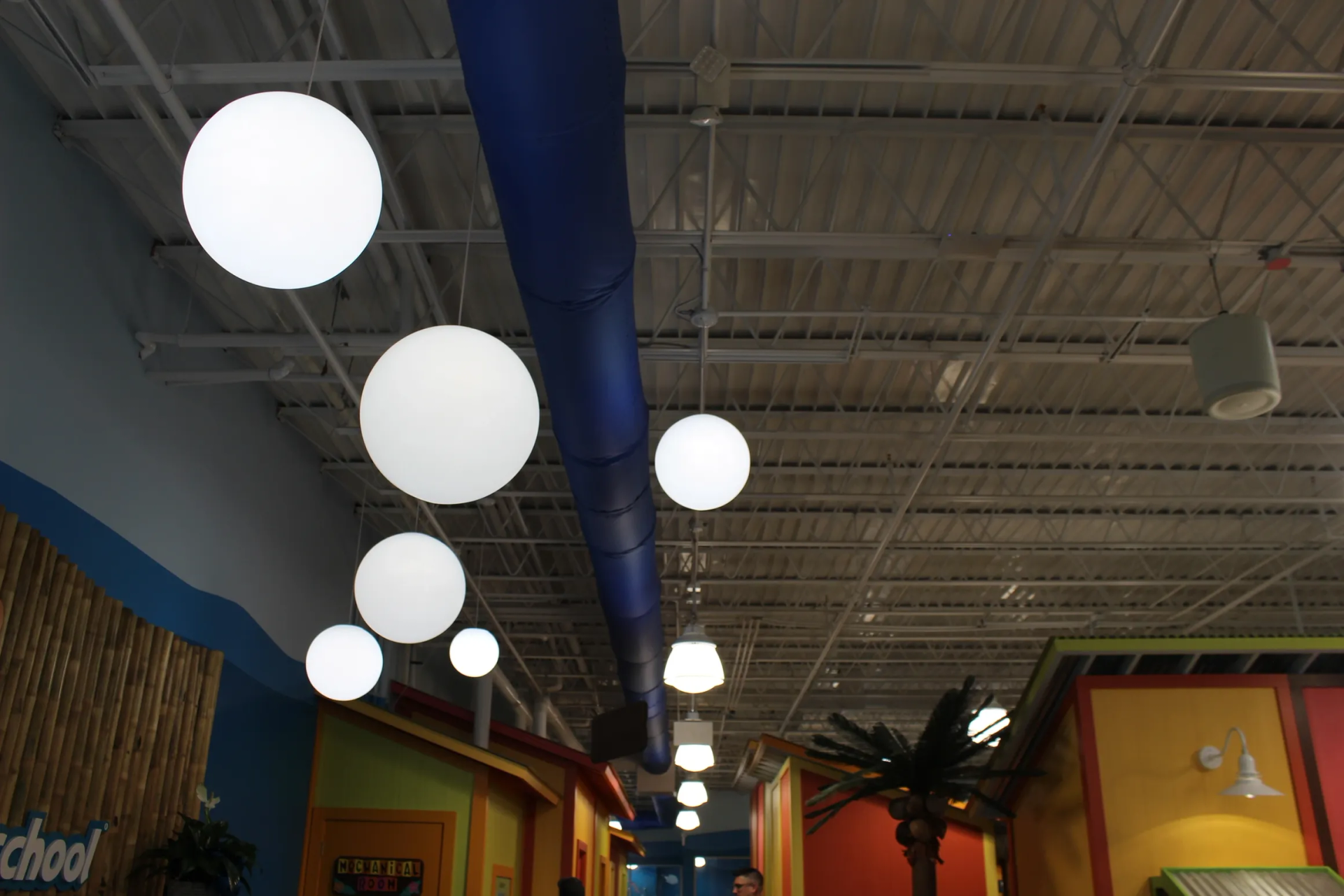 White globe lights hanging from a white ceiling with a blue duct, leading into a colorful corridor.