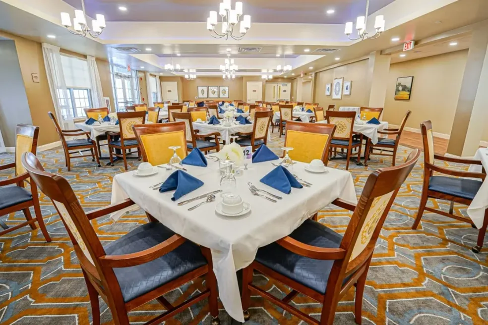 Dining room with tables set for a meal. Blue napkins and chairs, white tablecloths, and floral centerpieces.