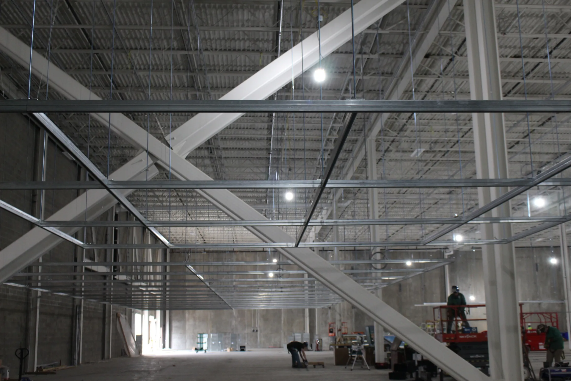 Interior of a large warehouse under construction, showing steel beams, girders, and ceiling framework. Workers on lift.