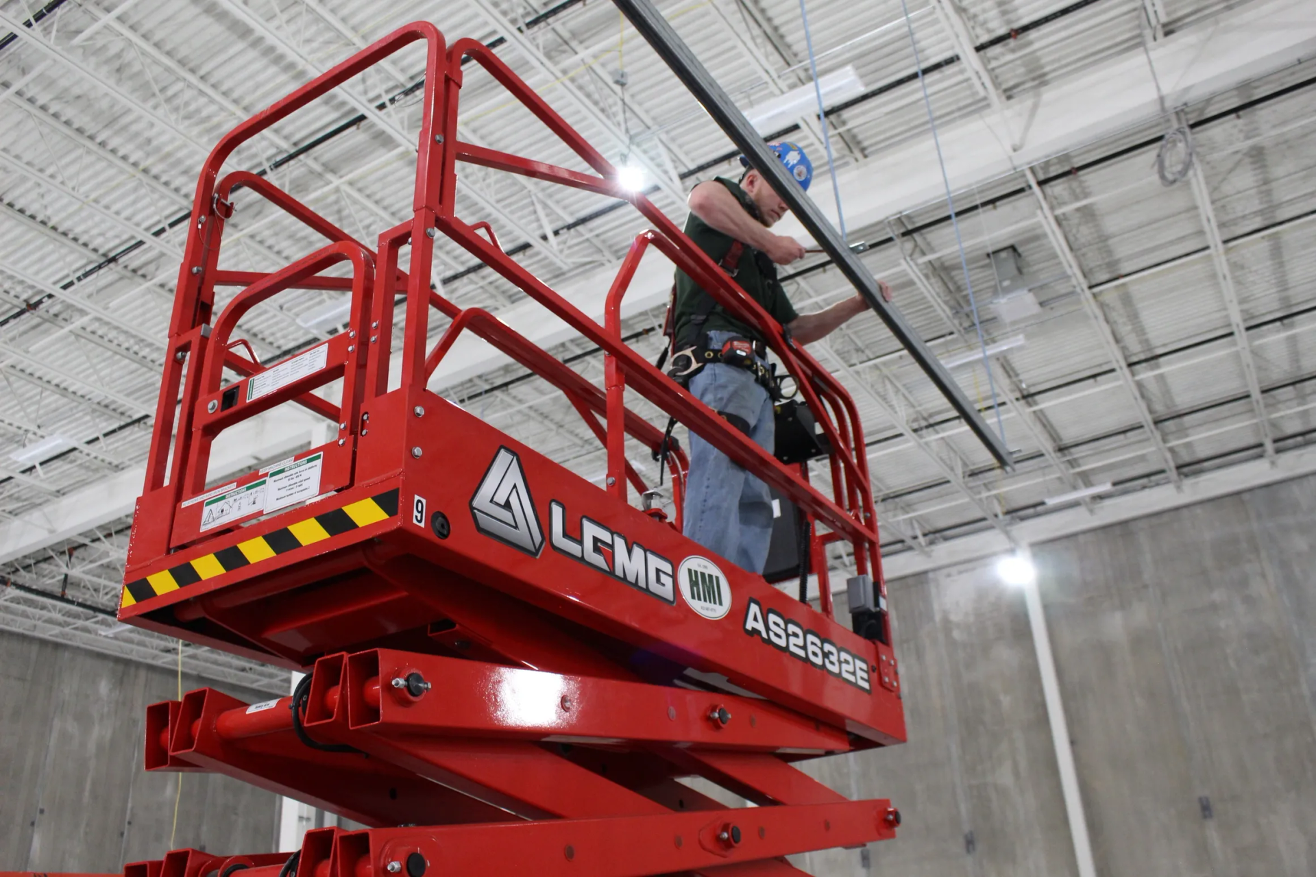 Worker in a red lift installing a metal bar on a ceiling, wearing a harness. Interior setting.