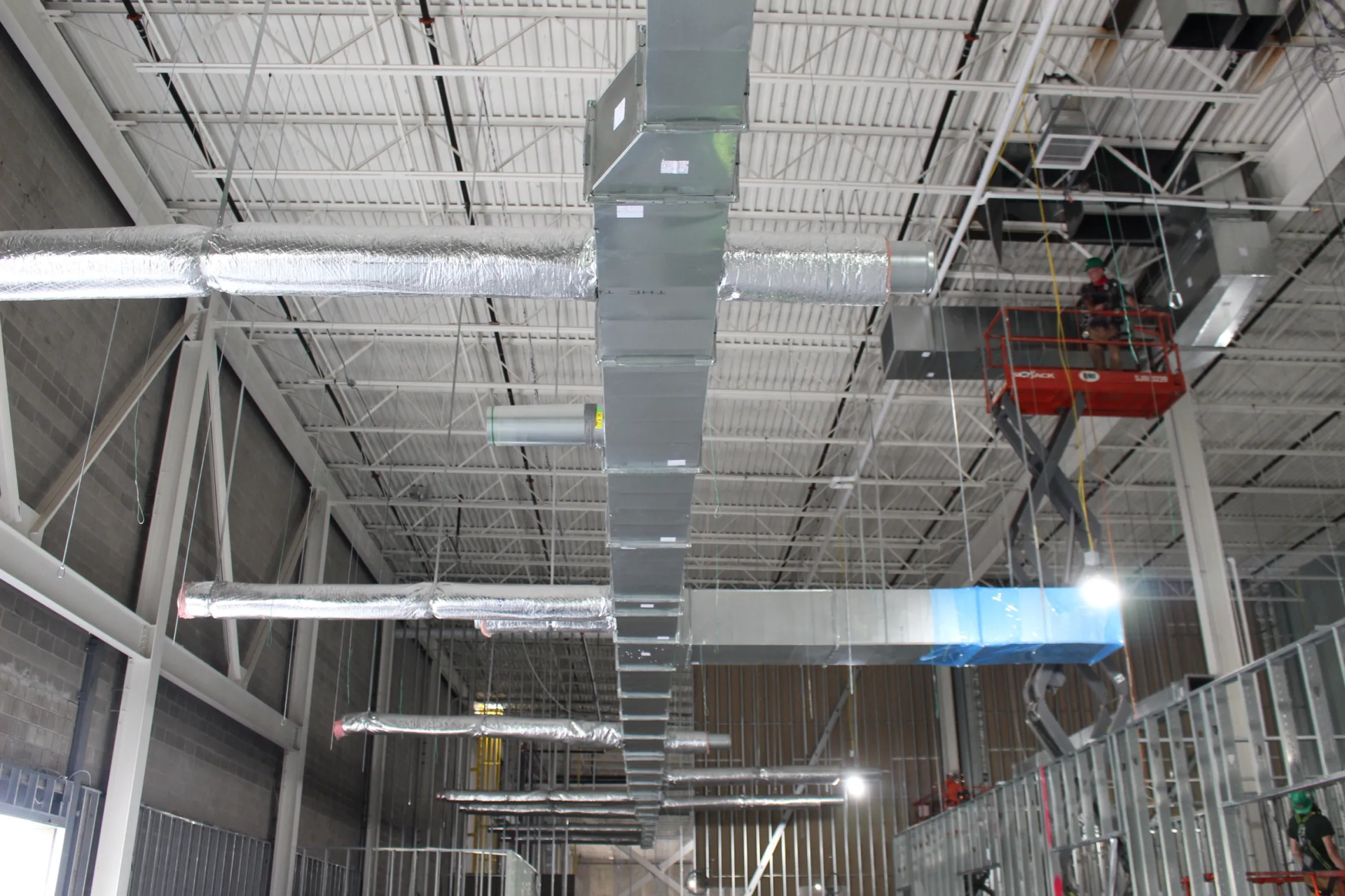 HVAC ductwork installation in a commercial space, with workers on a lift. Silver ducts run along the white ceiling.