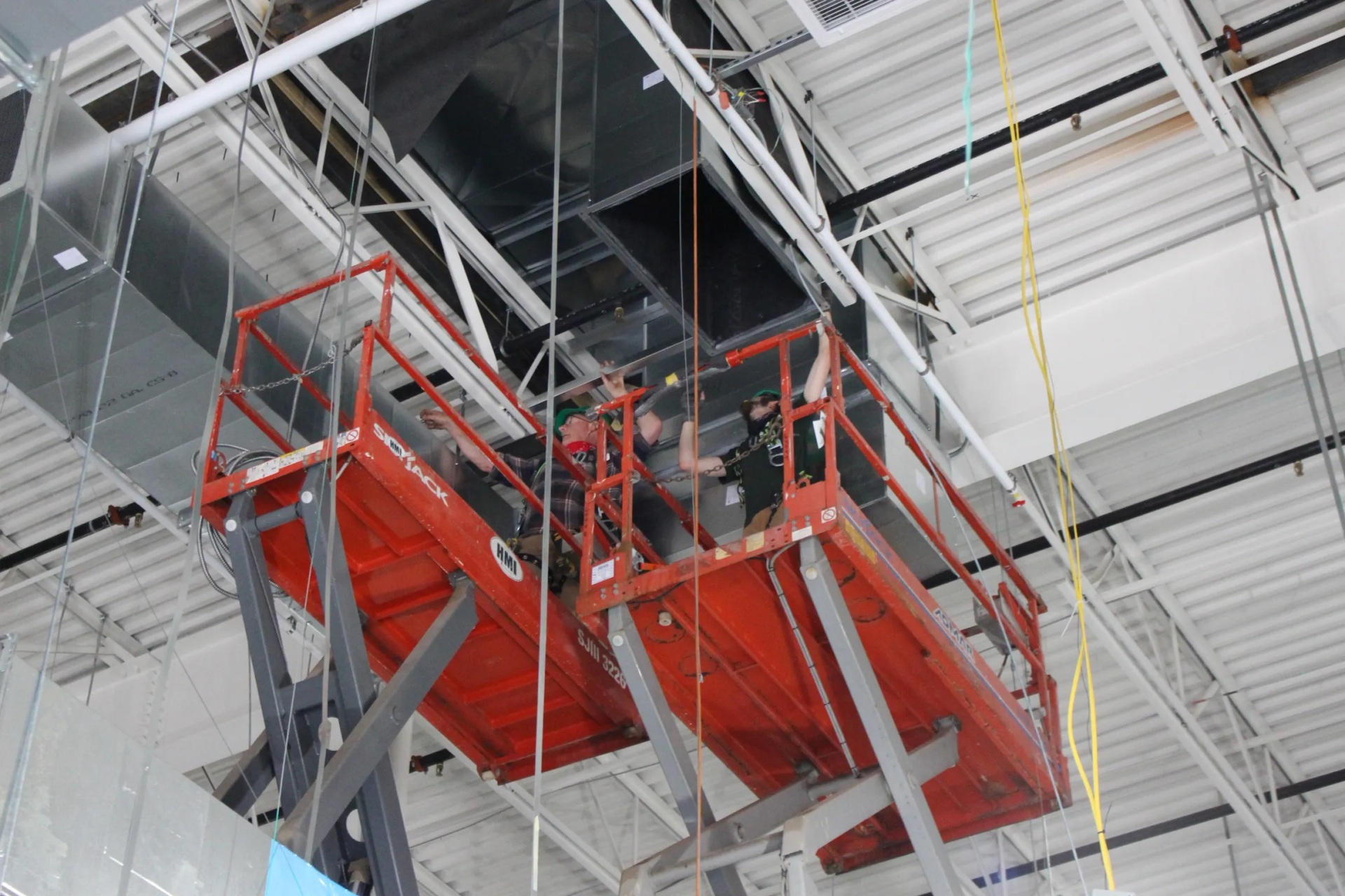 Orange scissor lift near ceiling, worker inside, working on ductwork.