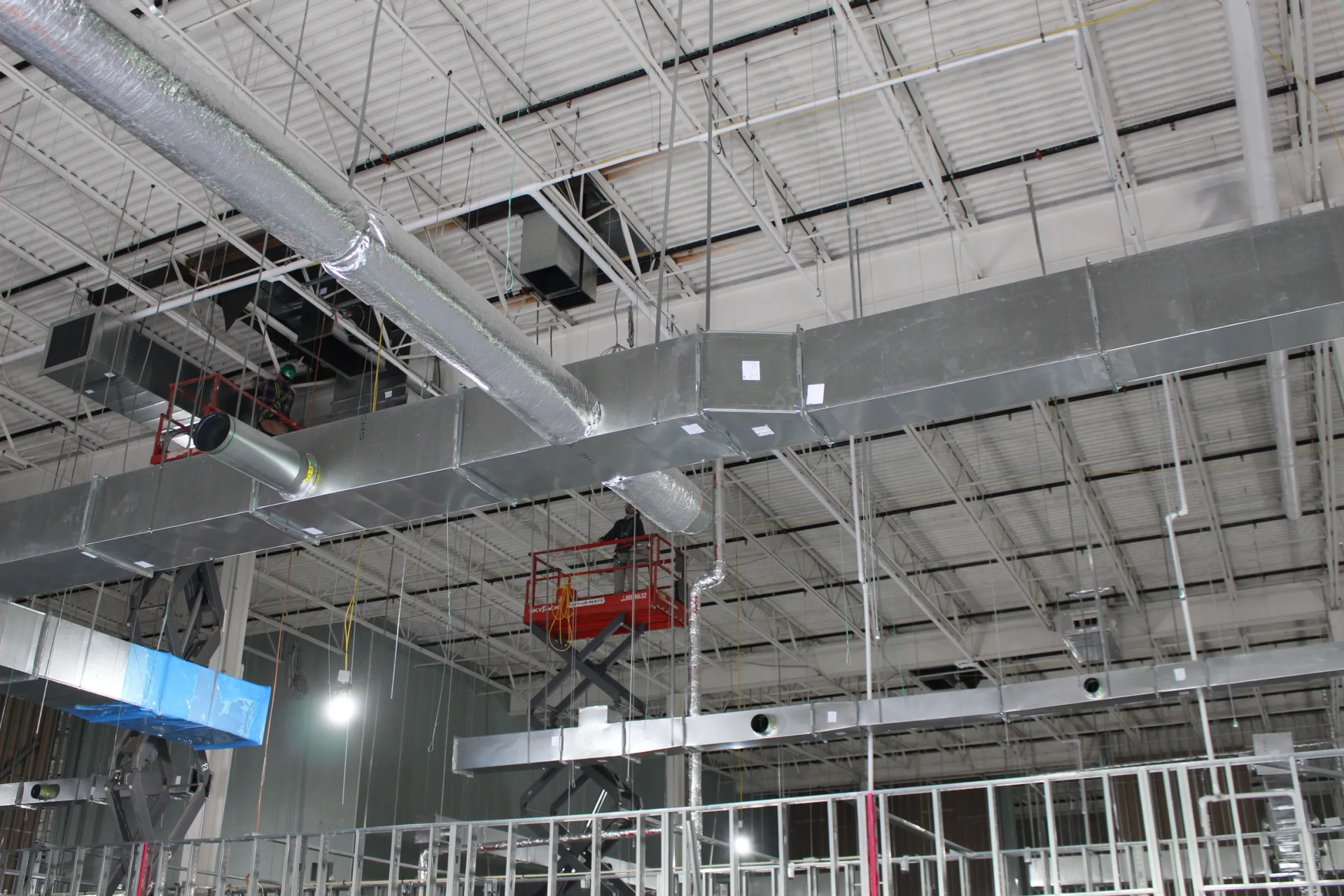 HVAC ductwork installation in a commercial building. A lift supports a worker near overhead metal ducts.