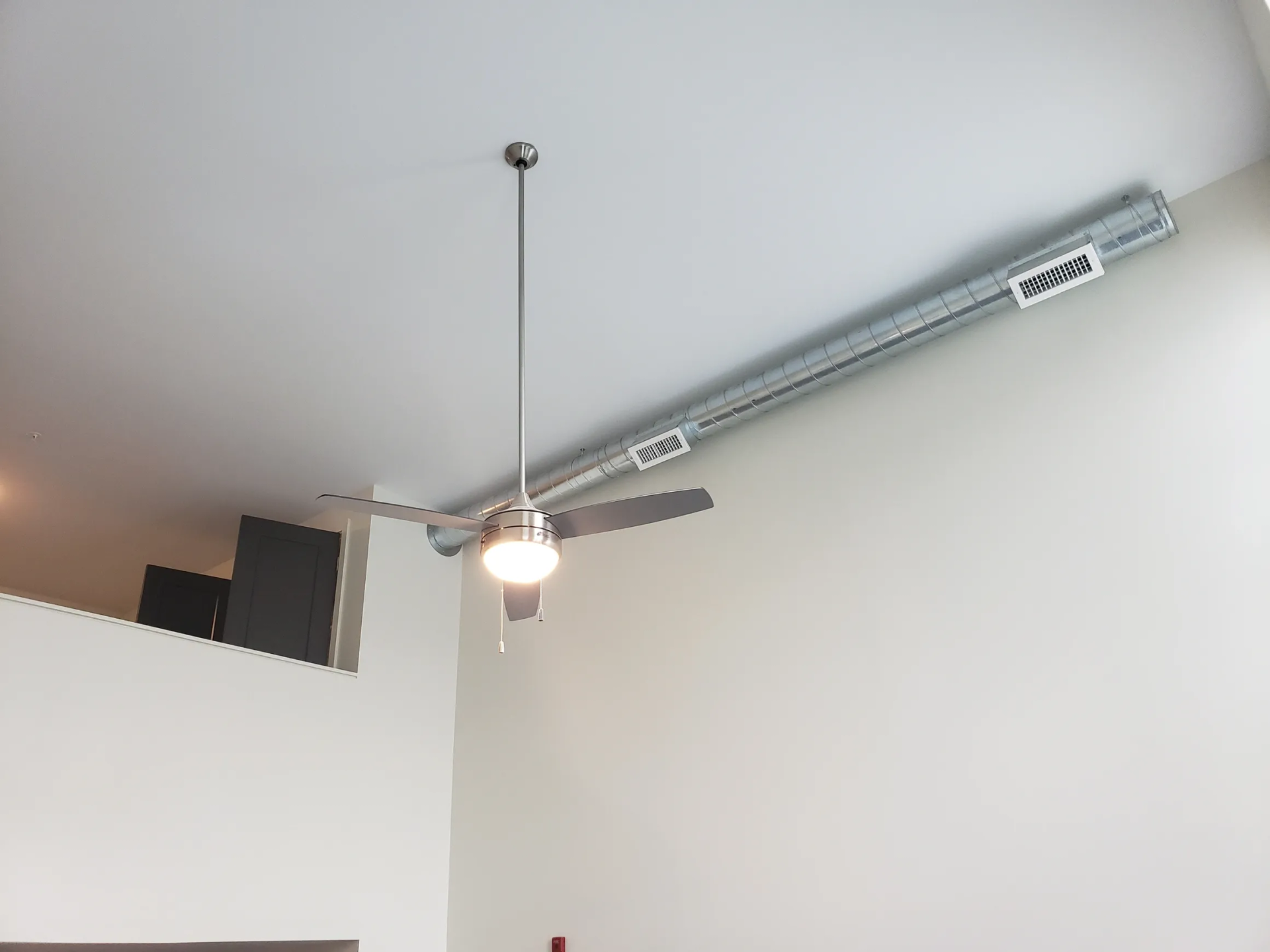 Ceiling fan with light fixture hanging from a high, slanted ceiling, next to a large metal duct.