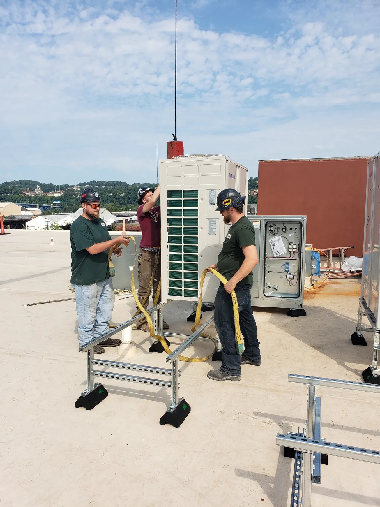 Three workers install equipment on a rooftop. One adjusts a cable; others secure the structure. A radio tower is visible.