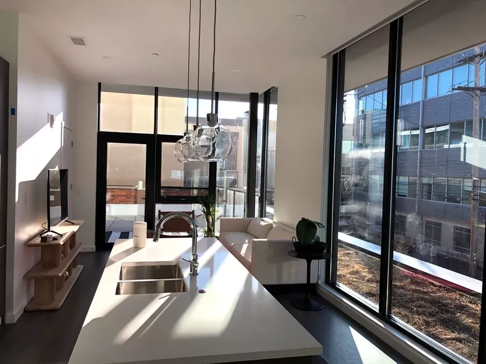 Modern, bright living space with kitchen island, white sofa, large windows, and outdoor balcony.