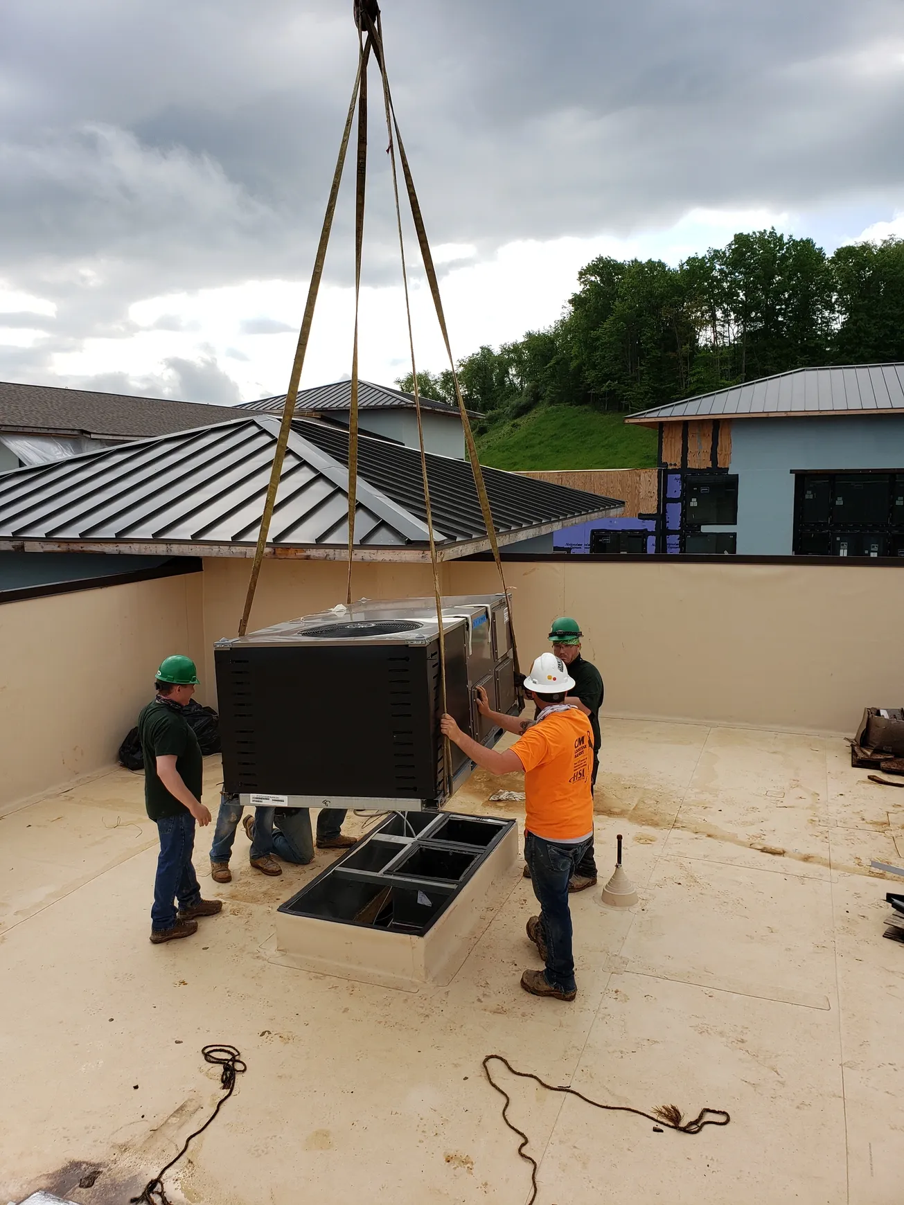 HVAC unit being lifted by crane onto a rooftop, workers in safety gear guiding it. Overcast sky.