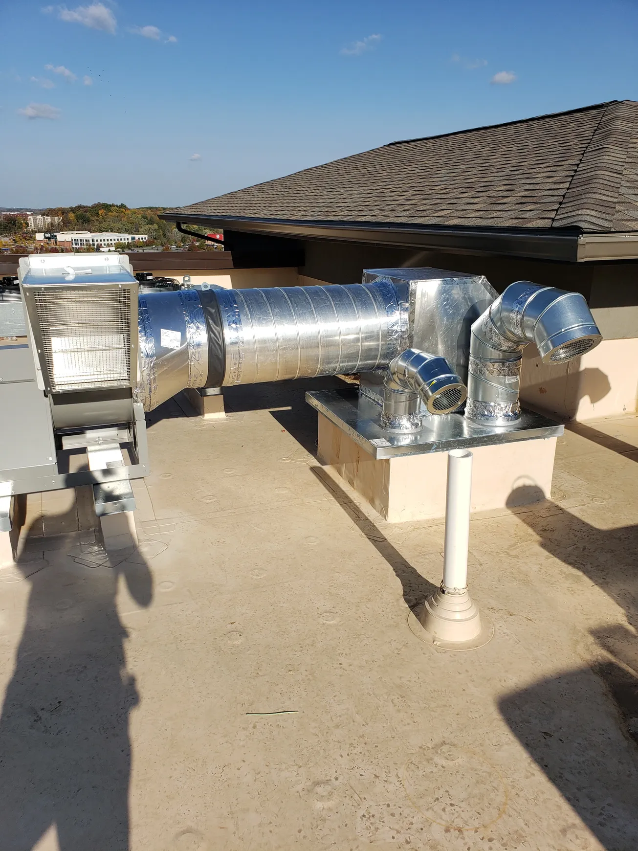 HVAC system on a rooftop. Metal ductwork, silver in color, connected to a rectangular enclosure and supported by a white pipe.