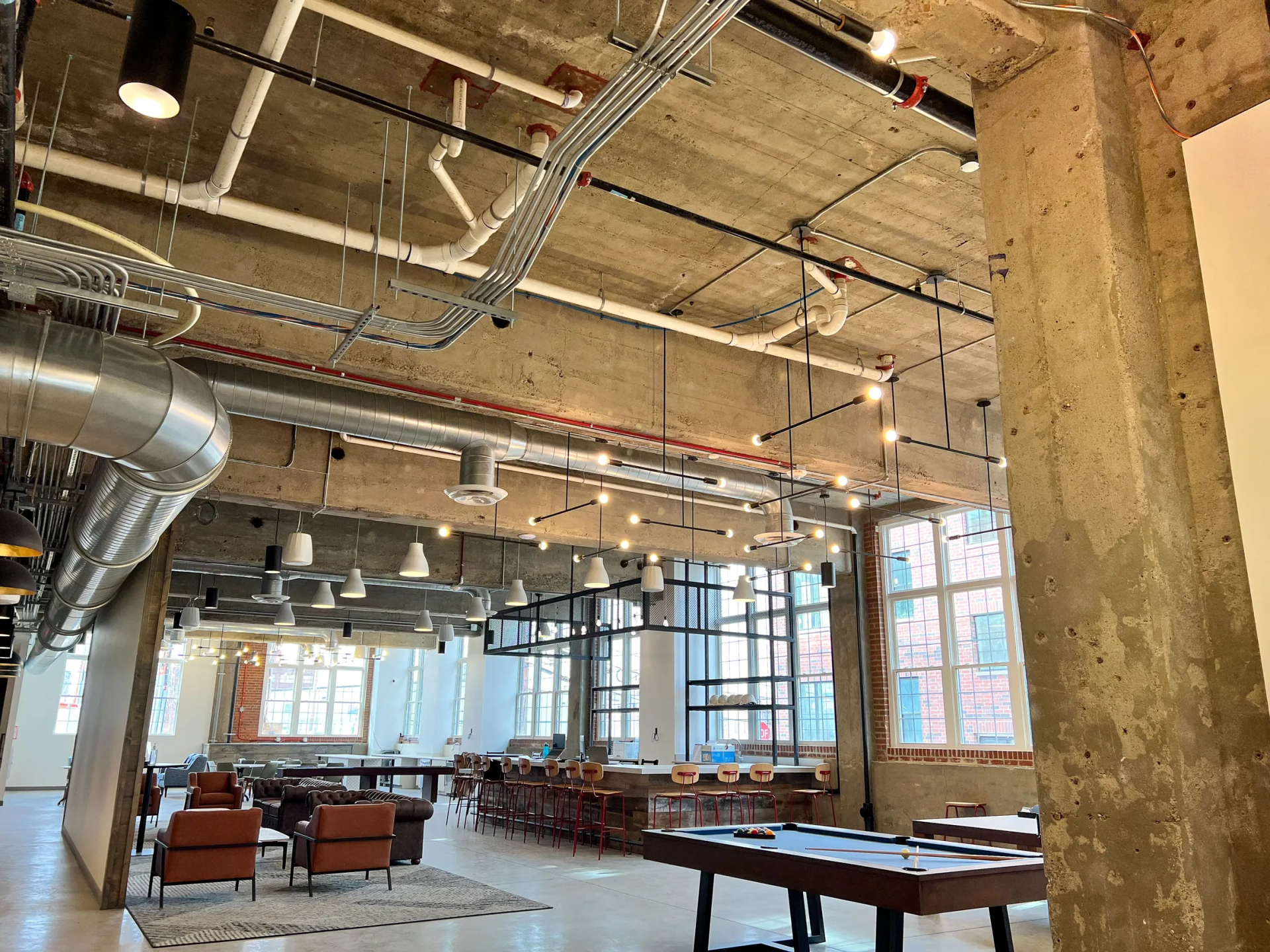 Open-plan office space with exposed concrete ceiling, industrial design, and many windows. Furnished with tables and chairs.