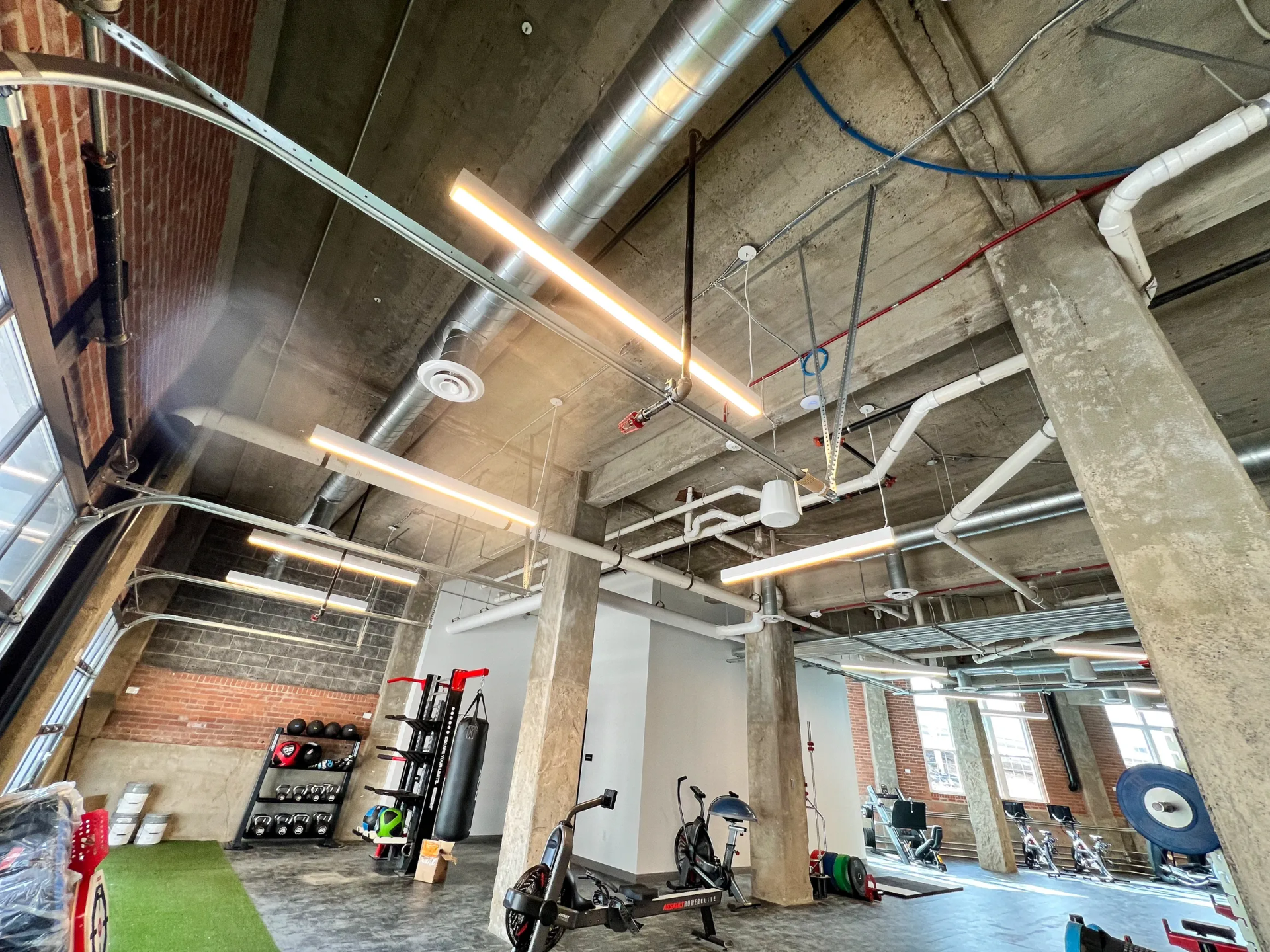Gym interior with exposed brick and concrete pillars, exercise equipment, and overhead lighting.
