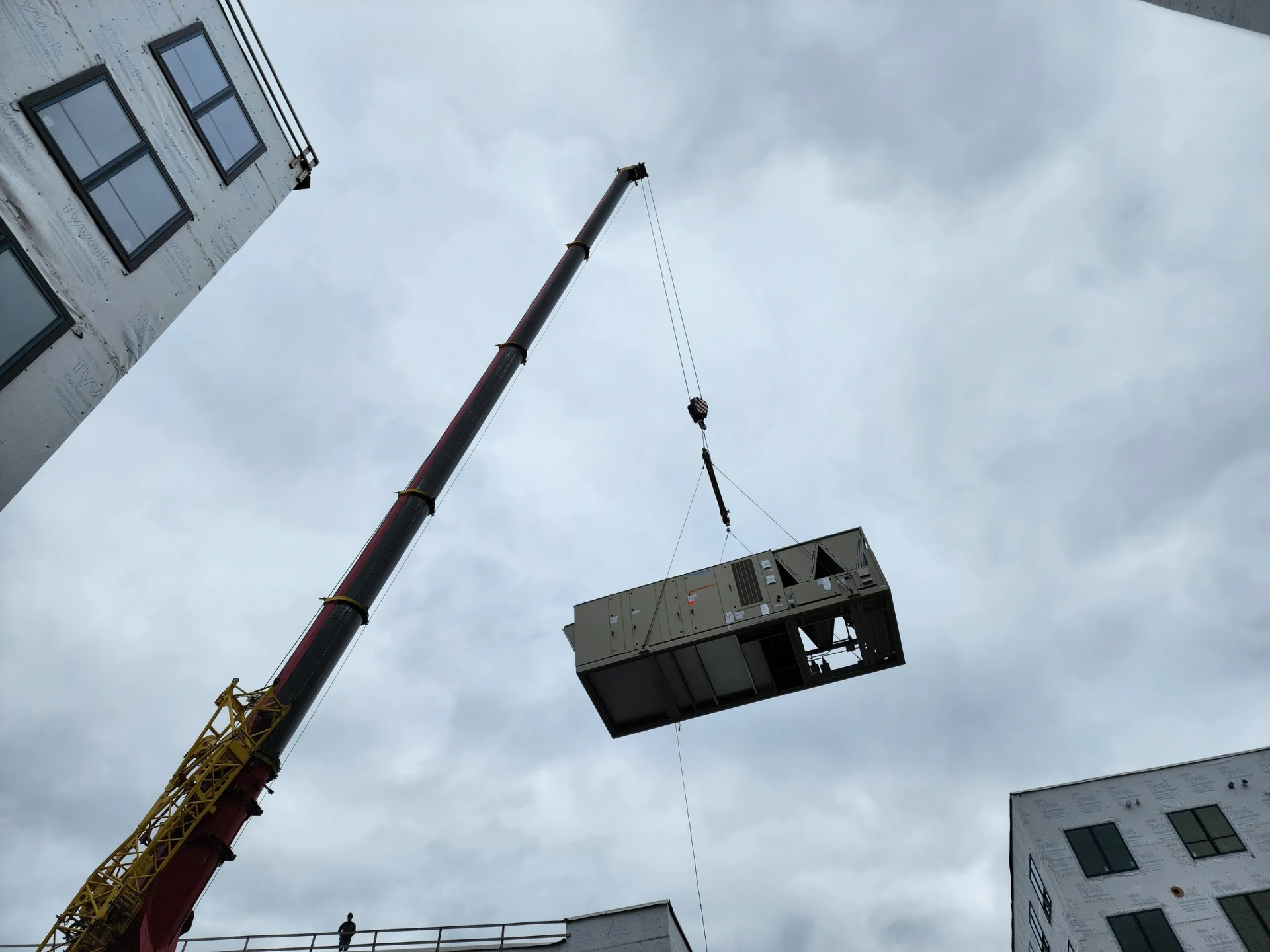 Crane lifting an HVAC unit on a construction site. Cloudy sky, two unfinished buildings.