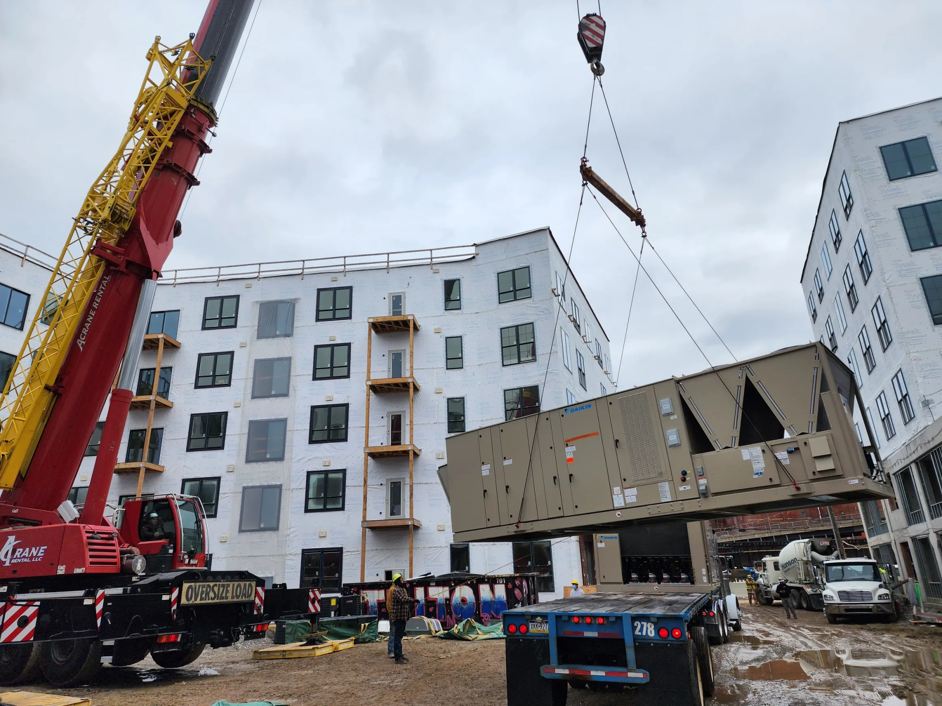 A crane lifting HVAC unit to the rooftop of a multi-story apartment building under construction.