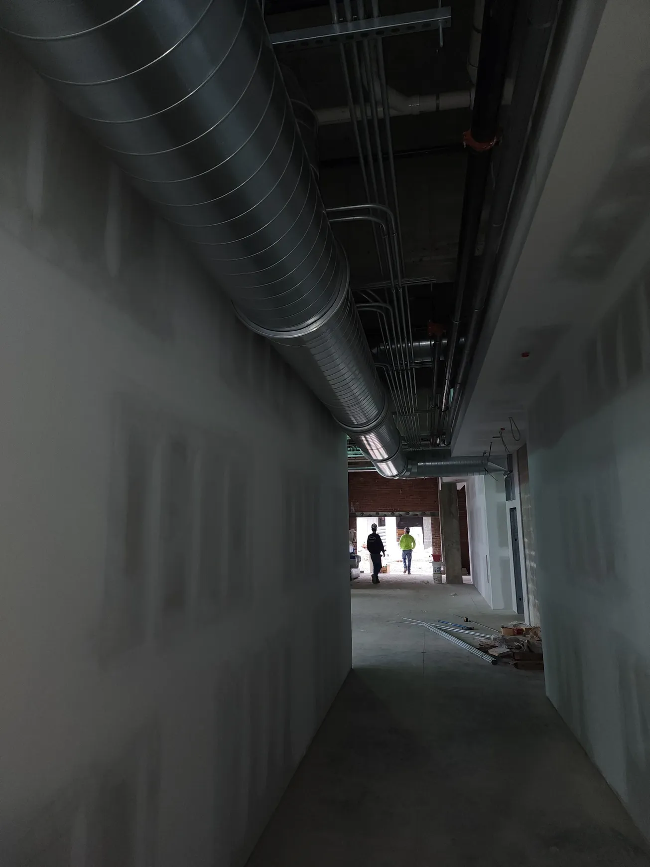 Narrow construction hallway with exposed ductwork, two figures at the end near a doorway.