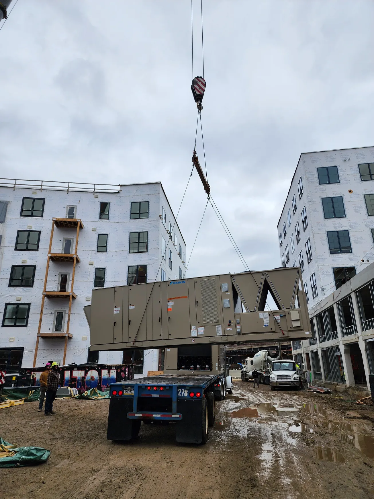 Crane lifting a large building panel near two apartment buildings under construction. Cloudy sky.