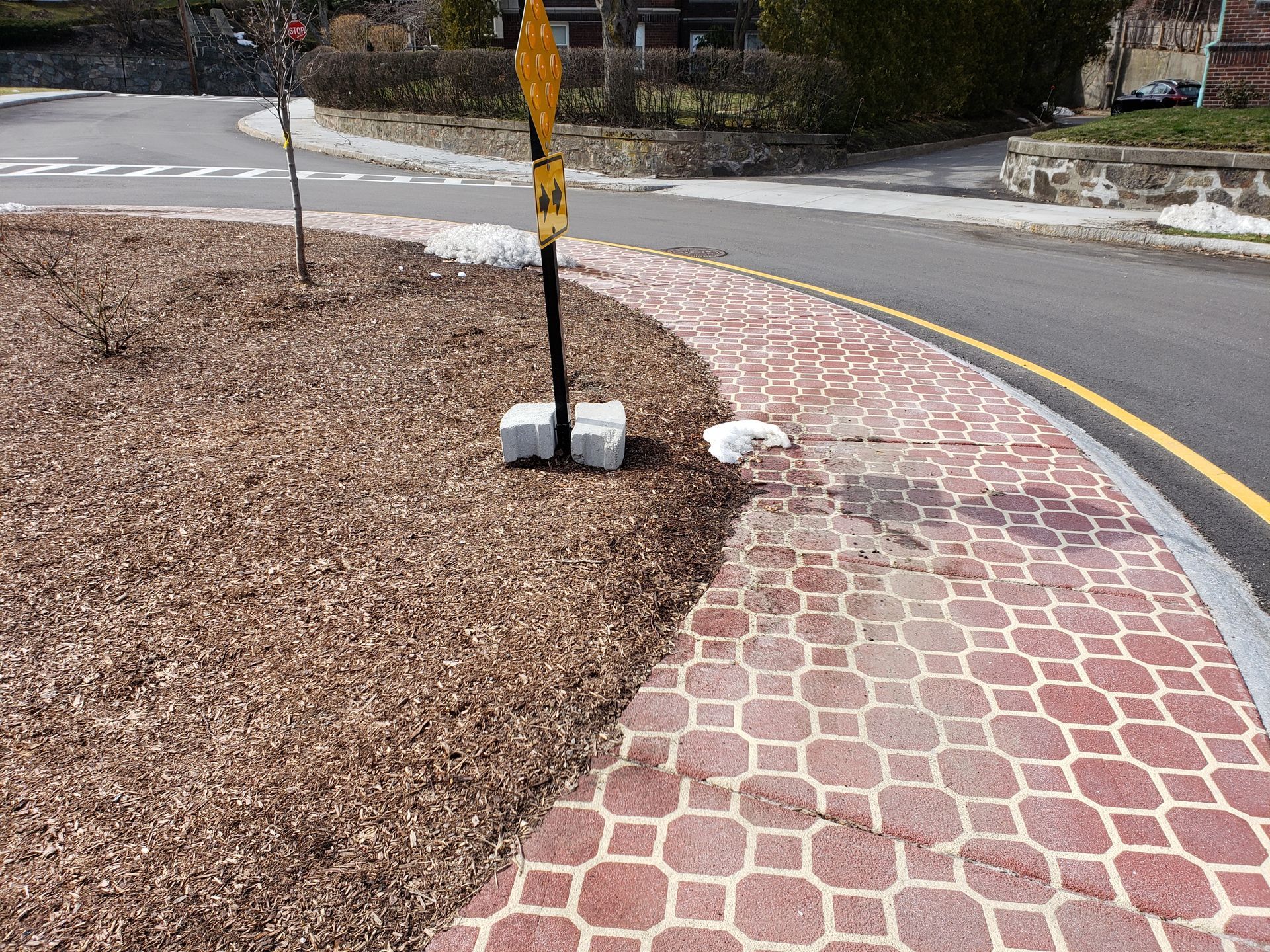 A yellow sign is sitting on the side of a road next to a brick walkway.