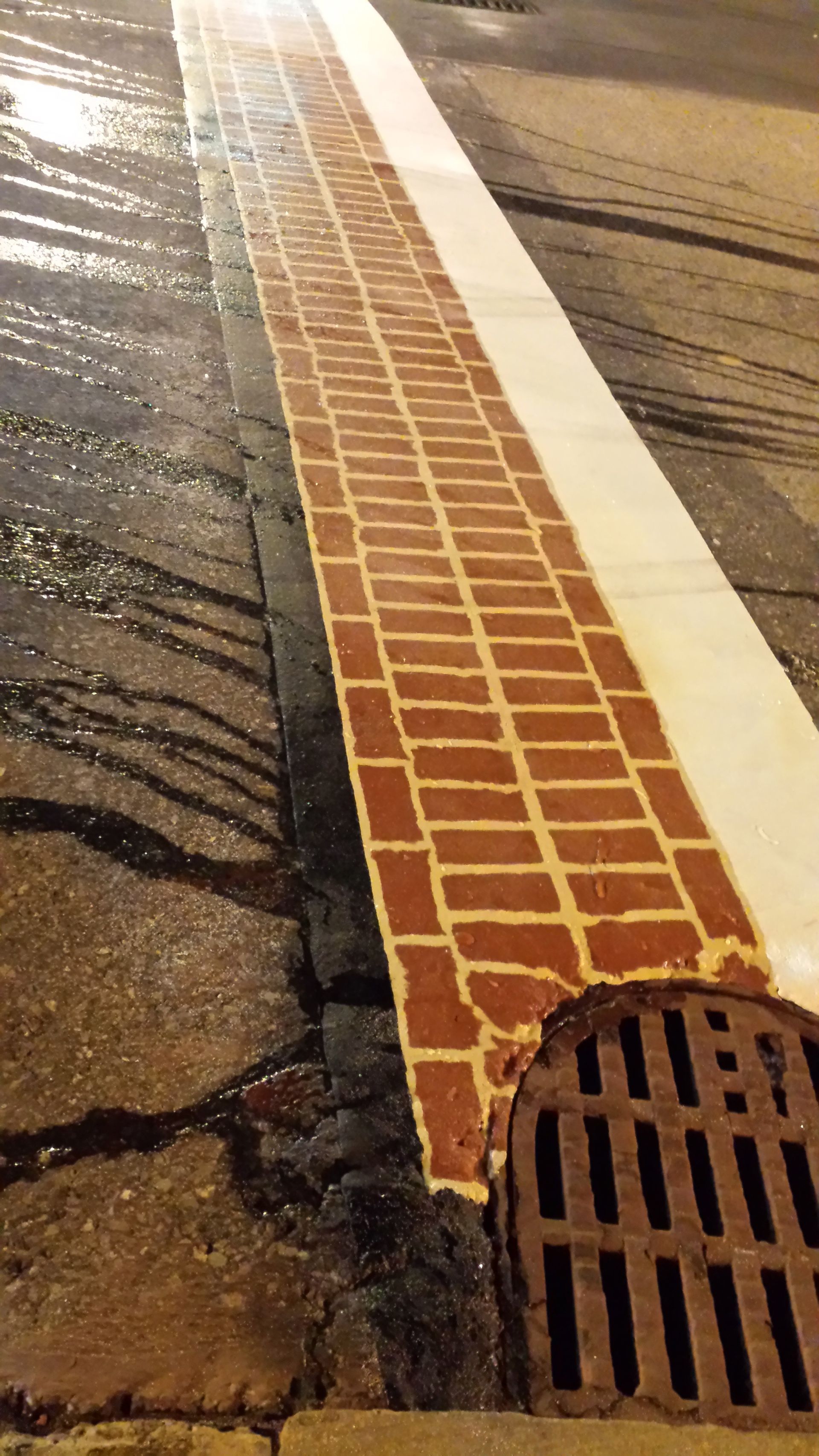 A drain on the side of a road next to a brick walkway.