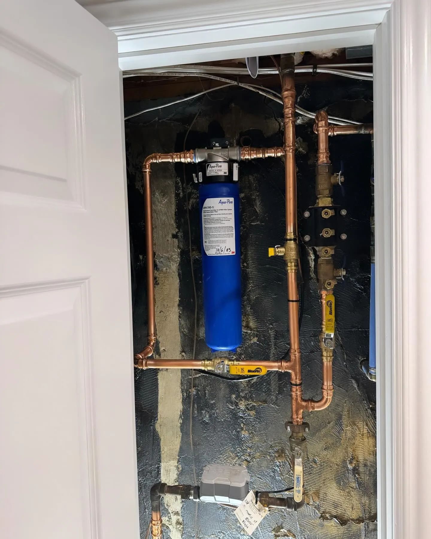 Open utility wall with exposed copper pipes and a blue water filter tank above a sink cabinet.