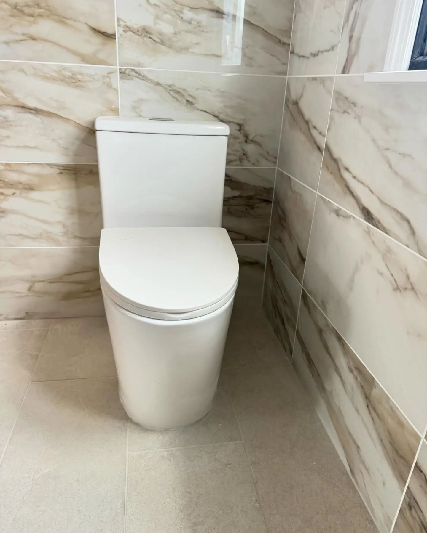 Modern white toilet in a tiled bathroom corner with beige marble-pattern walls and light floor.