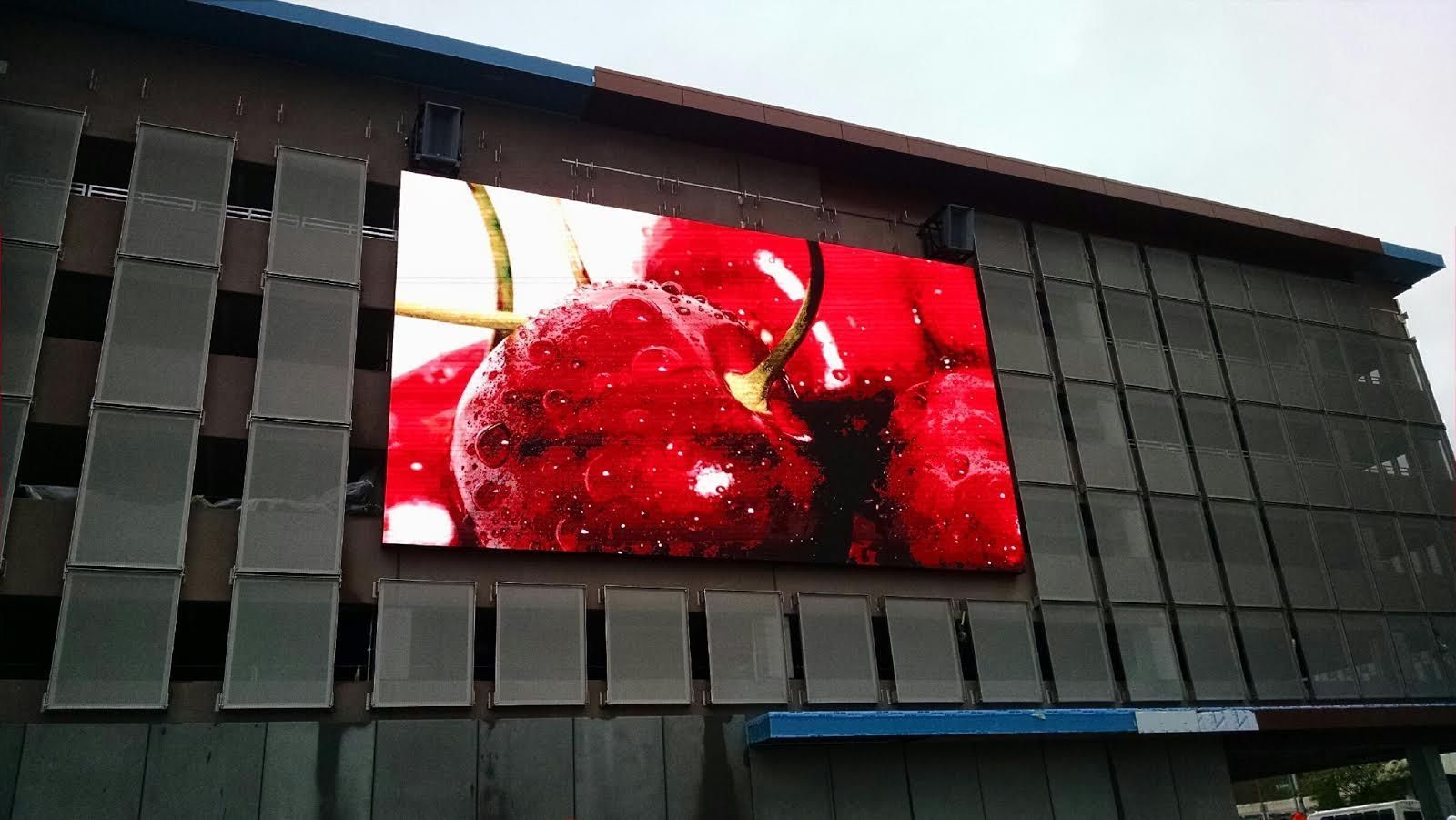 Large outdoor digital display shows close-up of ripe cherries; mounted on a building exterior with dark gray slats.