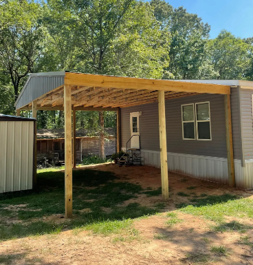 Wooden carport attached to a small house with a covered patio and trees in the background