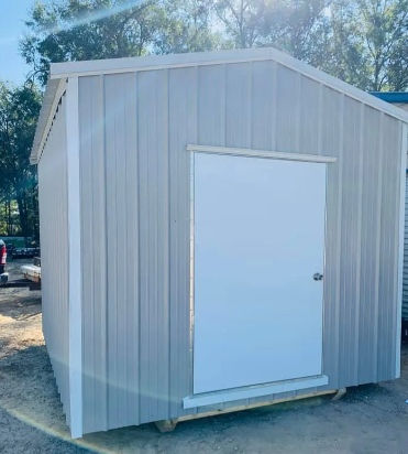 Small beige shed with a white door on a dirt lot, surrounded by trees.