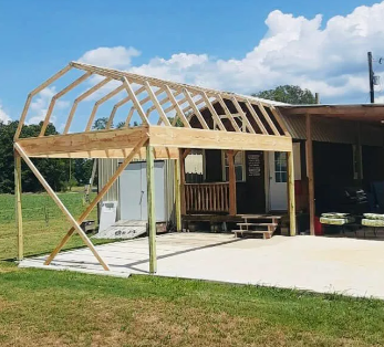 Partially built wooden carport frame beside a small house with a concrete pad and grassy yard