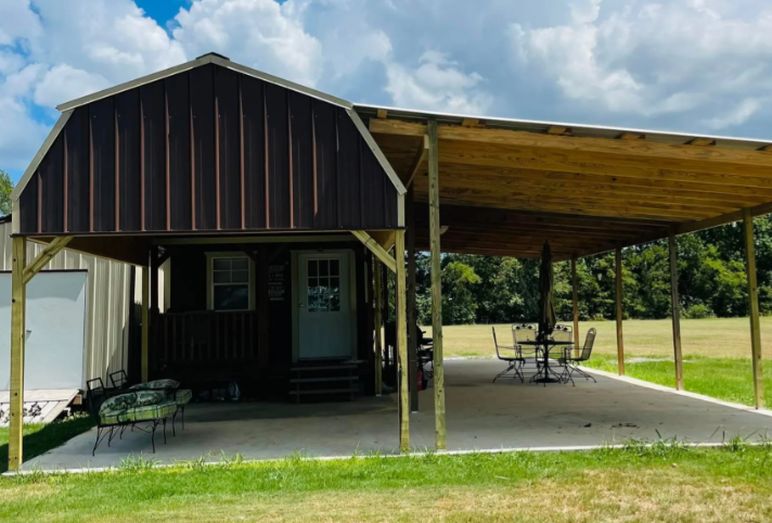 Small rustic cabin with covered porch and carport on a grassy lot under a cloudy sky