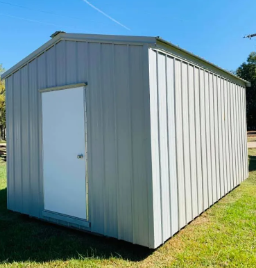 Gray metal storage shed with a white door on a grassy lot under a blue sky