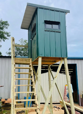 Small teal elevated shed on wooden stilts with ladder, under construction beside a metal building