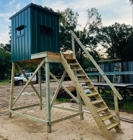 Elevated teal hunting blind on wooden stilts with stairs in a sandy outdoor clearing