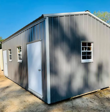 Gray metal storage shed with white doors and windows on a dirt lot in daylight