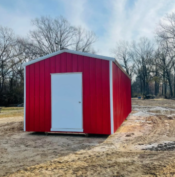 Red metal shed with a white door on a dirt lot, surrounded by leafless trees.