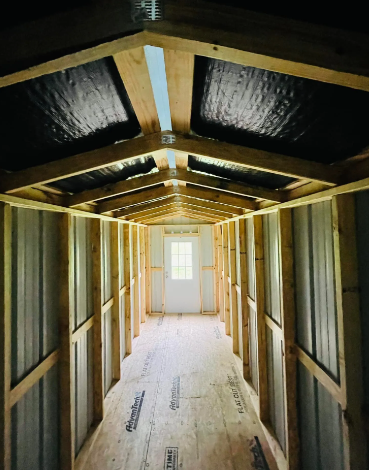 Narrow unfinished hallway with exposed wooden framing, insulation, and a bright door at the end.
