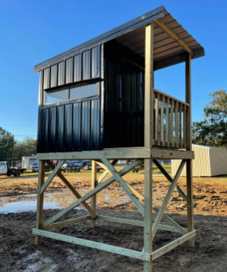 Small elevated black metal cabin on a wooden frame in a dirt lot, with a porch and slanted roof