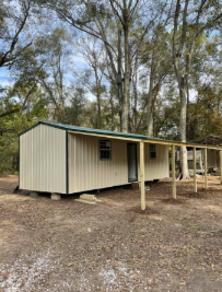 Small beige cabin with porch in a wooded area, with tall trees and dirt ground around it