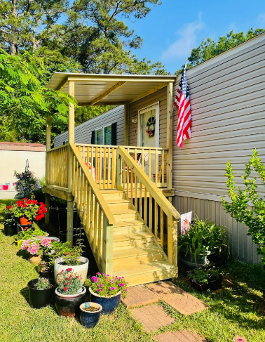 Front steps to a beige house with a covered porch, American flag, and potted flowers in a sunny garden.