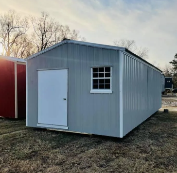 Small gray shed with white door and window, standing on grass near a red building at sunset
