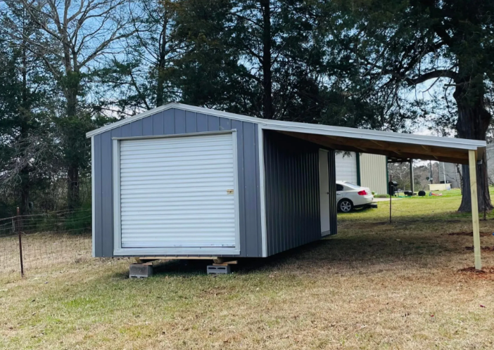 Gray shed-style carport with roll-up door, set on a grassy yard near trees and a parked car.