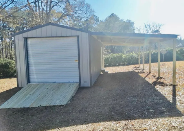 Small metal storage shed with roll-up door and ramp, beside a covered carport on a gravel lot.