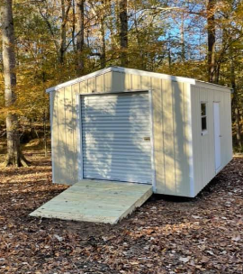 Small beige shed with a ramp and roll-up door in a wooded autumn setting