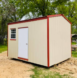 Small beige shed with red trim and a white door, standing outdoors on dirt near trees.