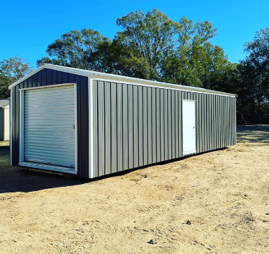 Gray metal storage shed with roll-up door and side entry door on a dirt lot under a blue sky
