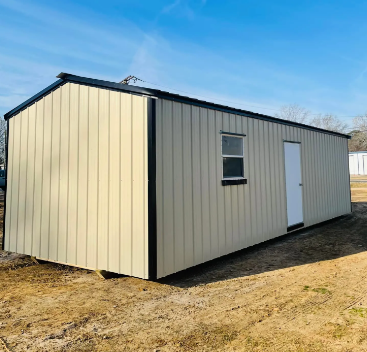 Small beige metal shed with black trim and a white door, sitting on bare ground under a blue sky